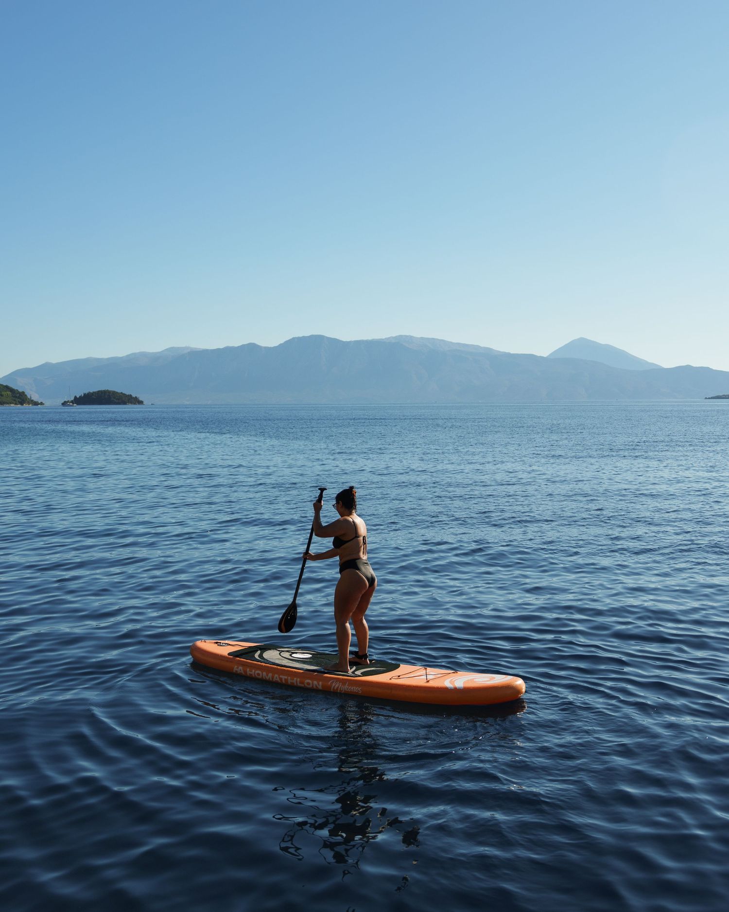 Paddleboarder glides across calm blue waters of a mountain lake with distant peaks on the horizon.