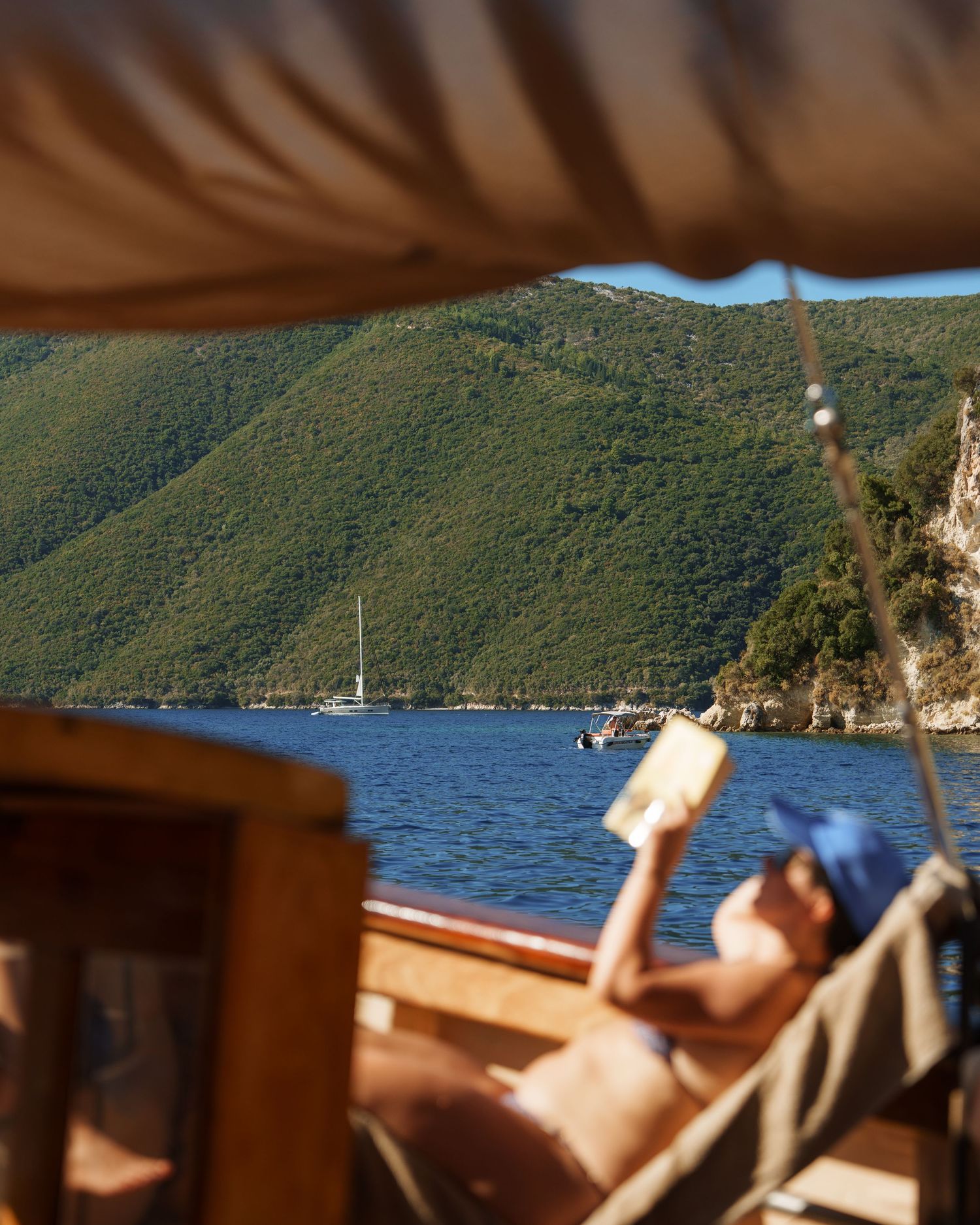 Person relaxing on a sailboat deck with mountain coastline and blue waters in the background.