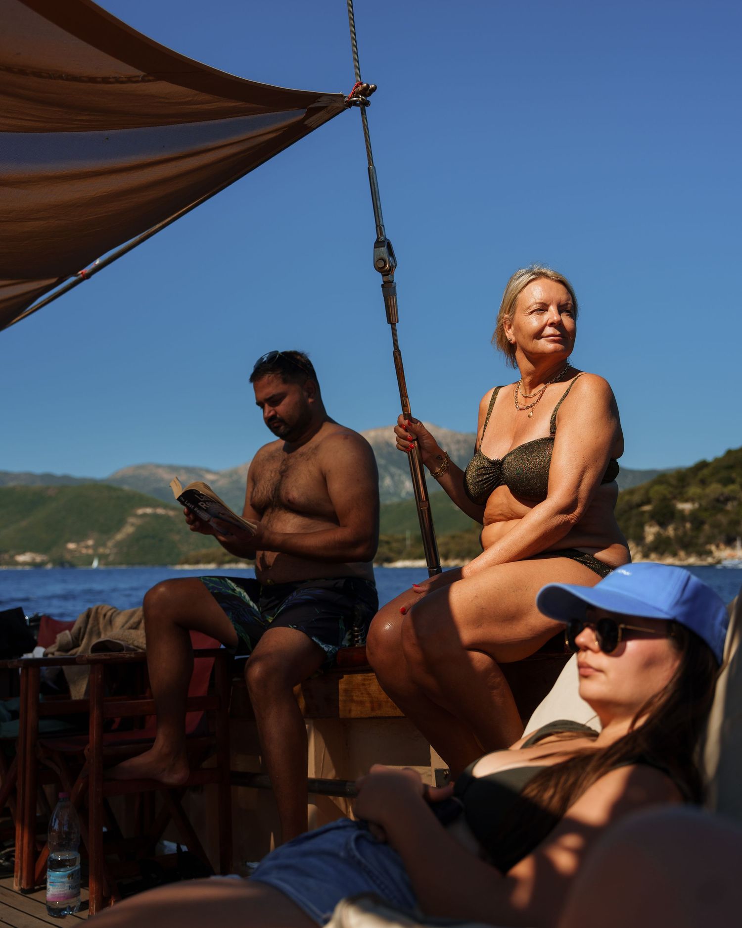 People relaxing under a brown sunshade on a boat deck with mountains and blue water in the background.