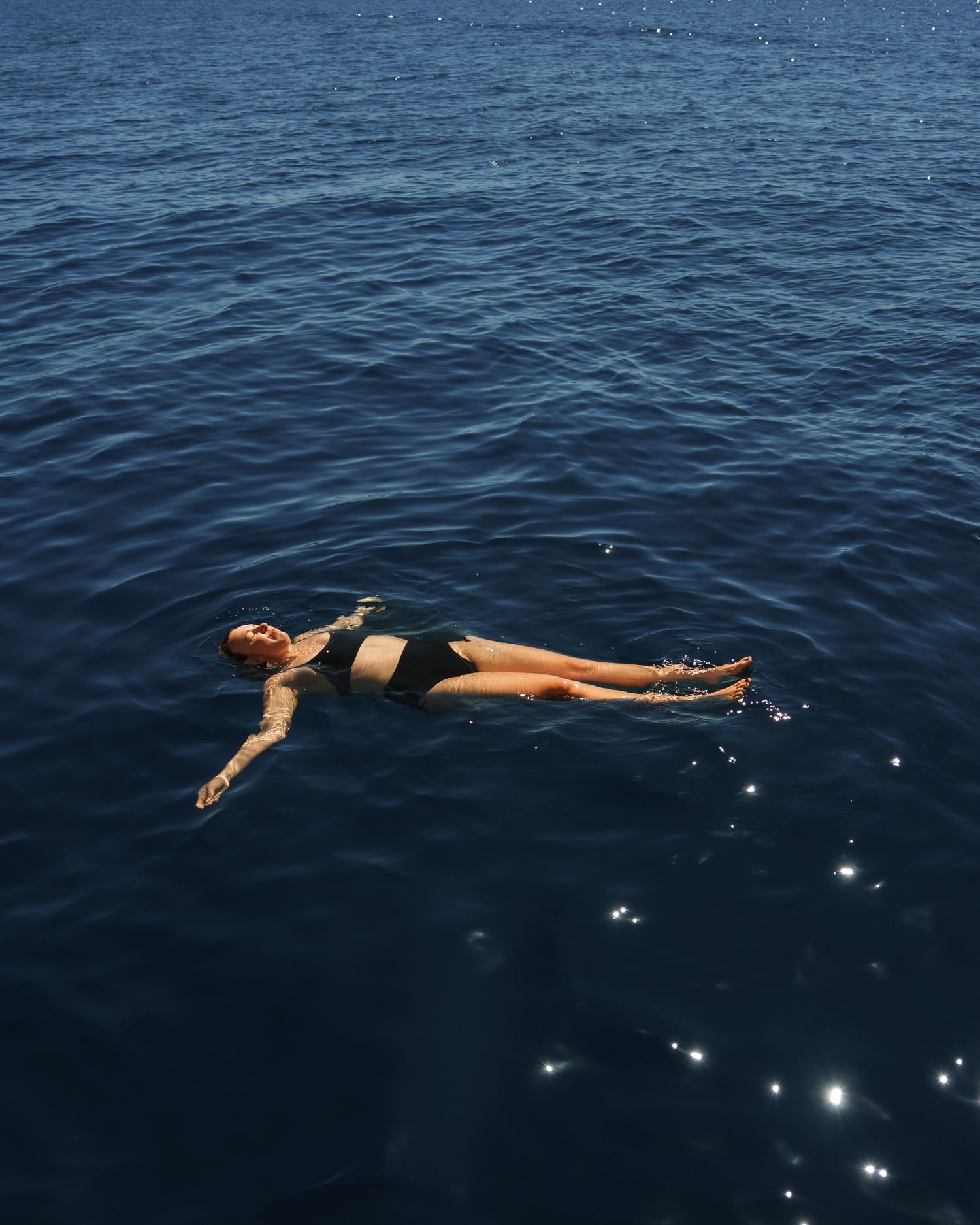 Person in black swimwear floating peacefully on calm blue ocean water with sunlight sparkling on the surface.