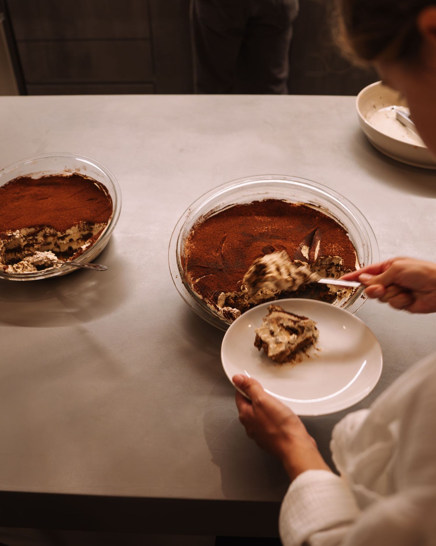 Hands serving a slice of chocolate dessert from a round pan onto a white plate with spoons and bowls on a table.