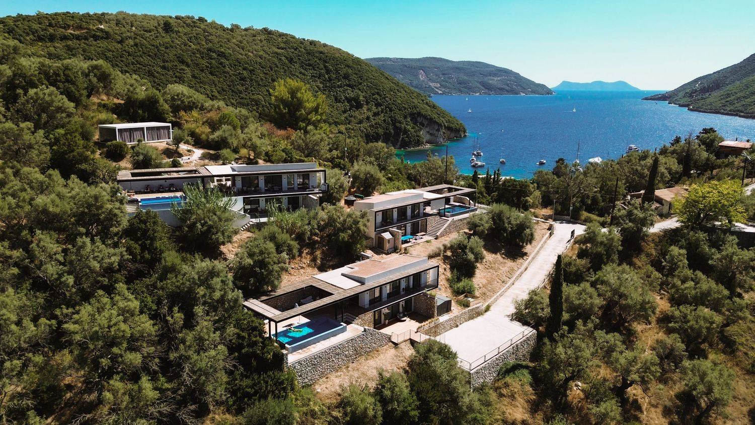 Panoramic view of a coastal landscape with modern hillside homes overlooking a blue bay surrounded by green mountains.