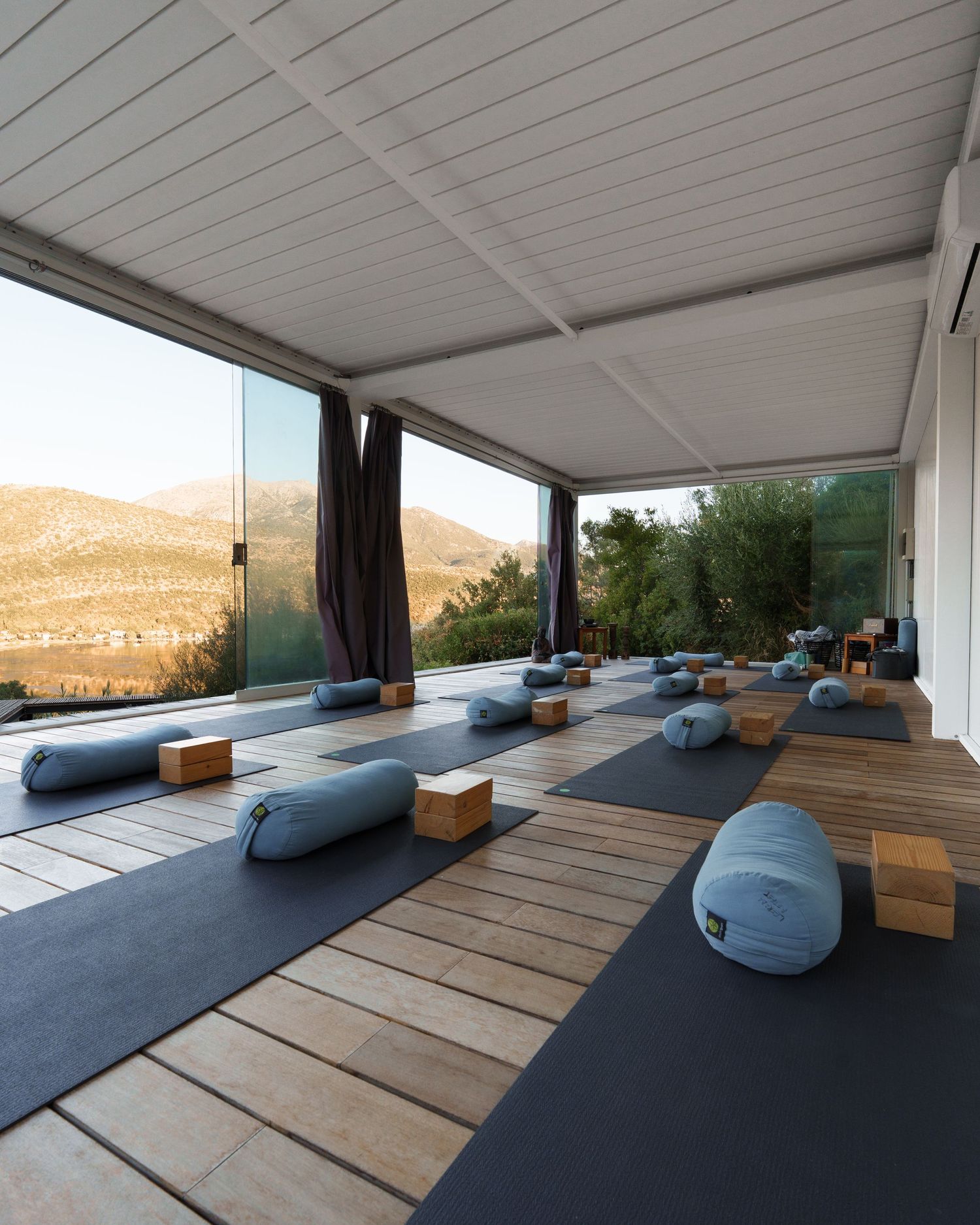 Modern yoga studio with blue cushions and wooden floors overlooking a scenic mountain landscape through large windows.
