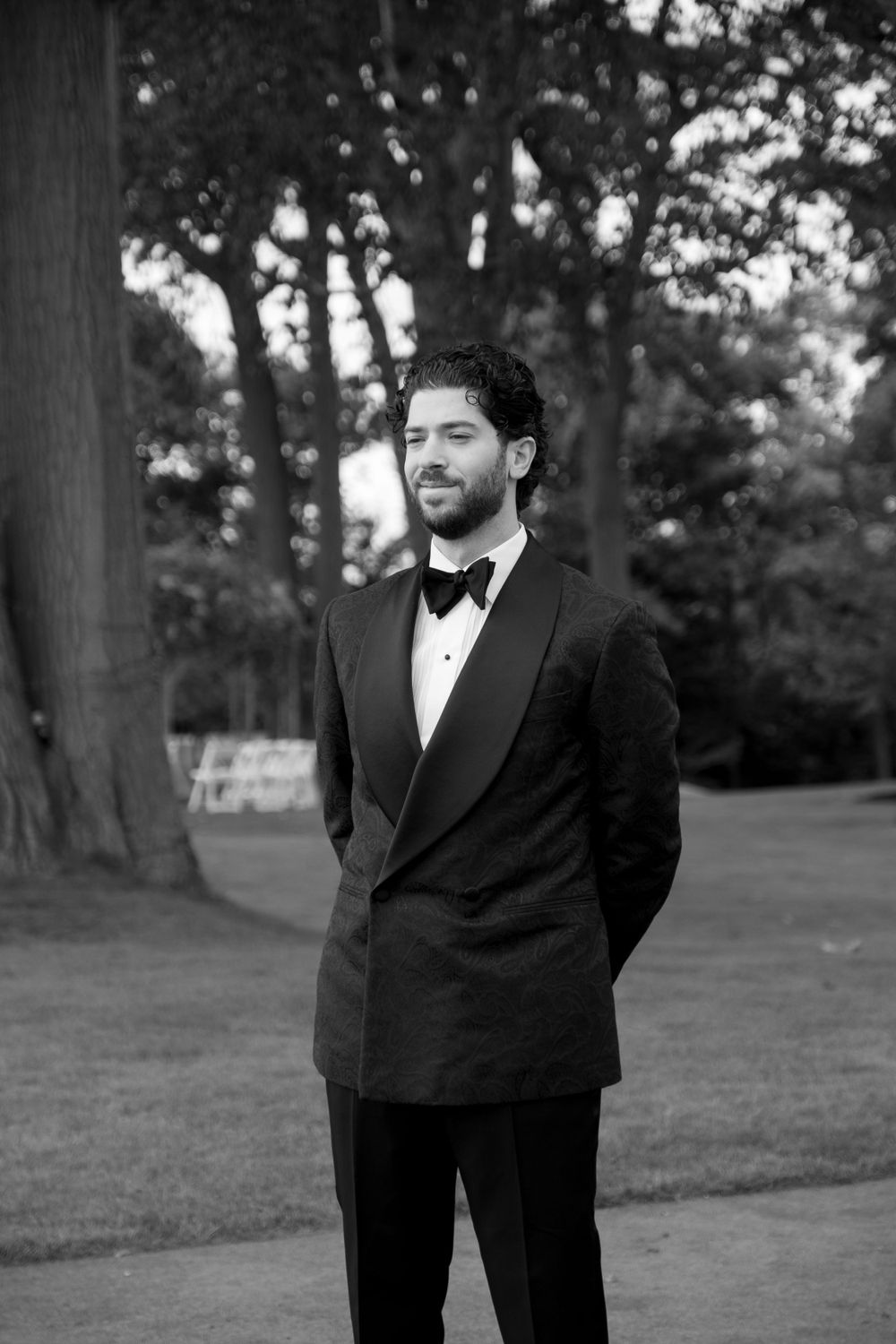 Black and white portrait of someone in a classic tuxedo with bow tie standing outdoors amid trees and grass.