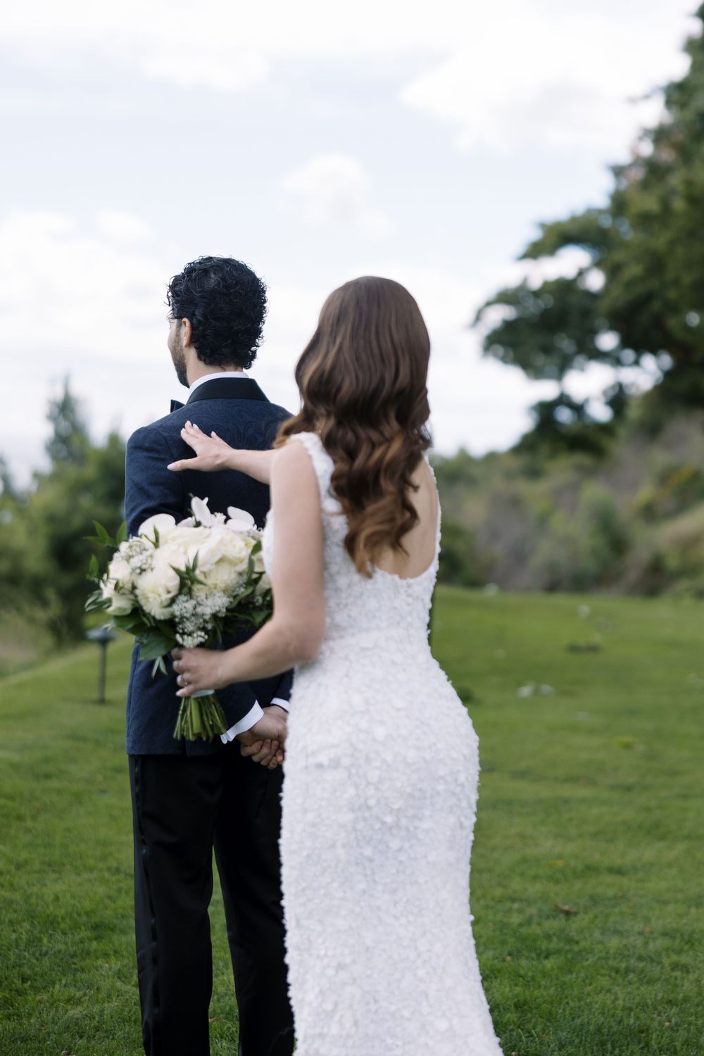 Newlywed couple in formal attire standing on green lawn with white rose bouquet looking toward trees.