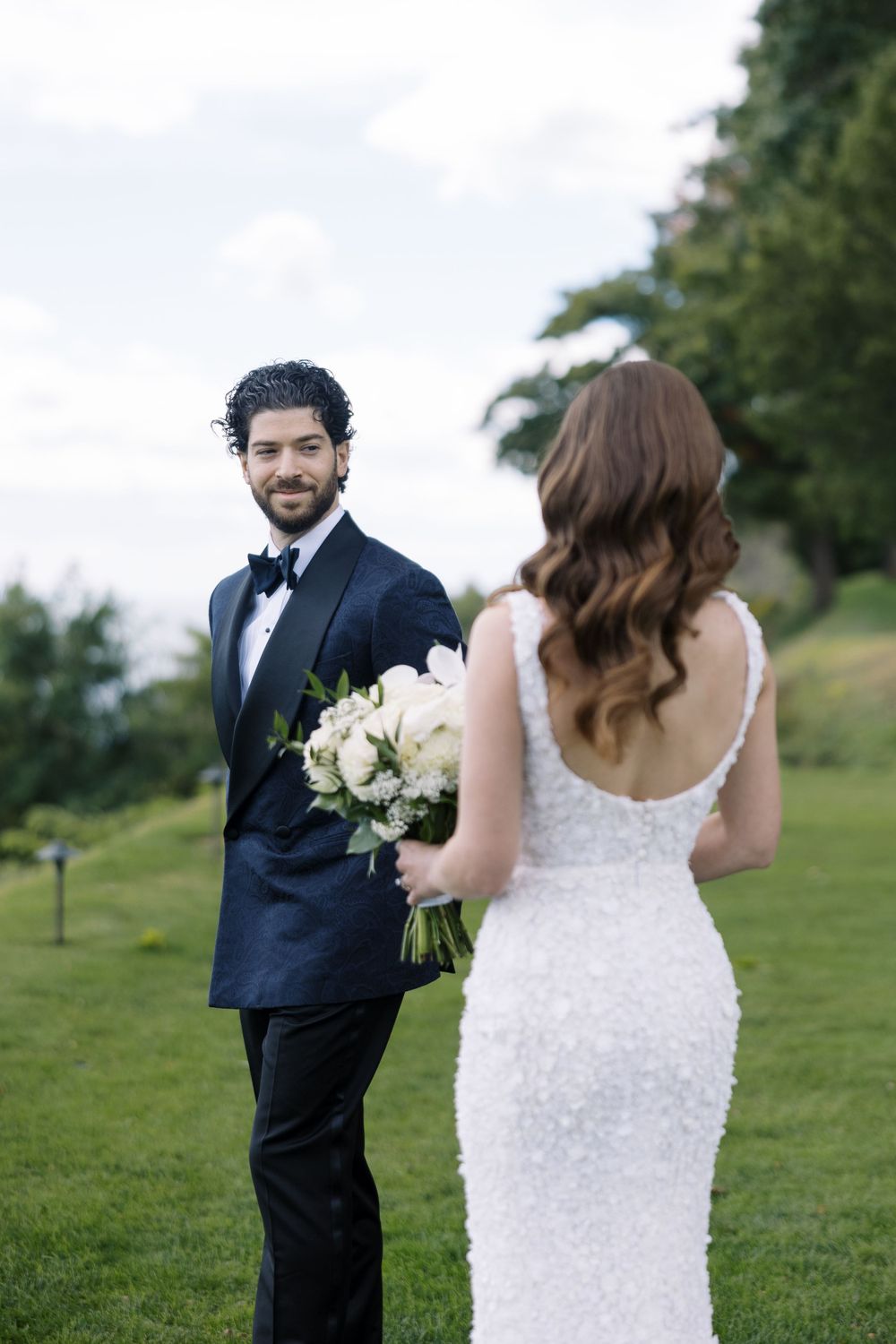 A couple in wedding attire stands on a grassy hillside, with the bride holding a white floral bouquet.