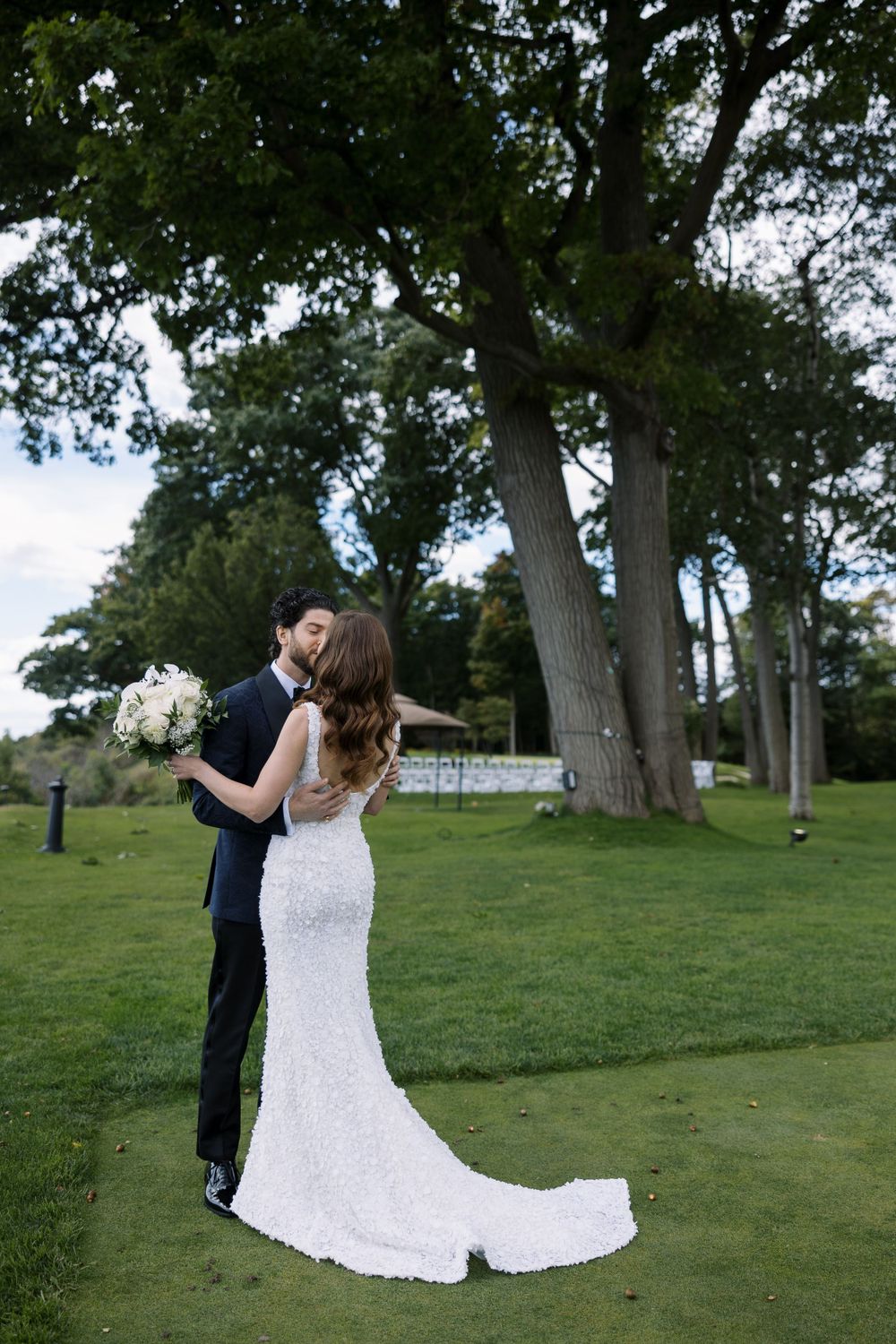 A couple shares a romantic moment on a lush lawn with majestic trees in the background, the bride wearing a fitted lace gown.