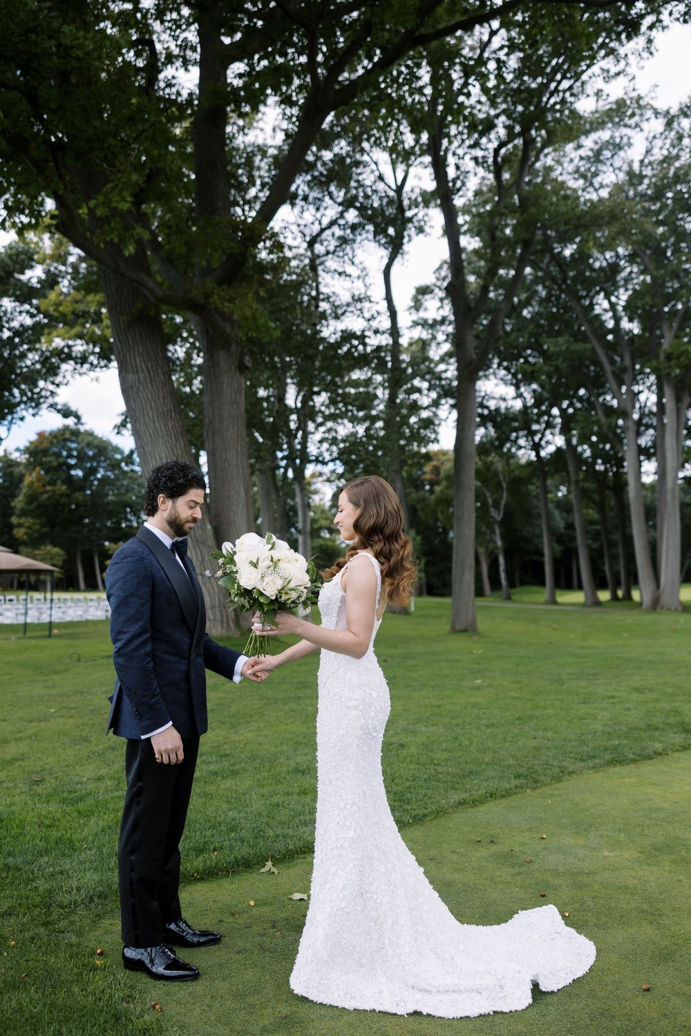A couple stands together during their wedding ceremony on a lush green lawn surrounded by tall trees.