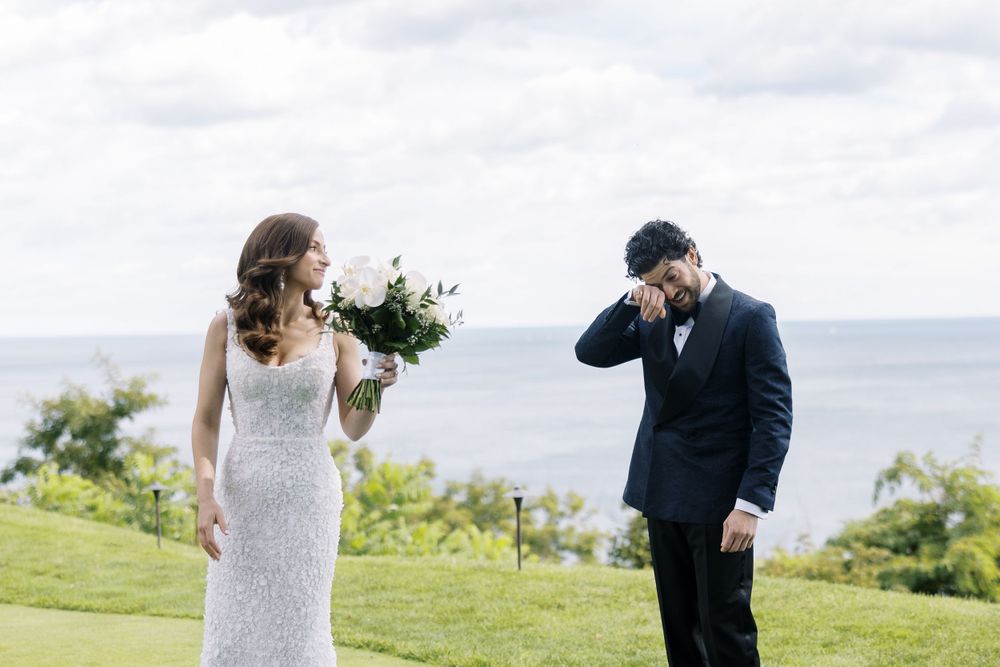 A joyful wedding moment captured outdoors on a green hilltop overlooking the ocean.