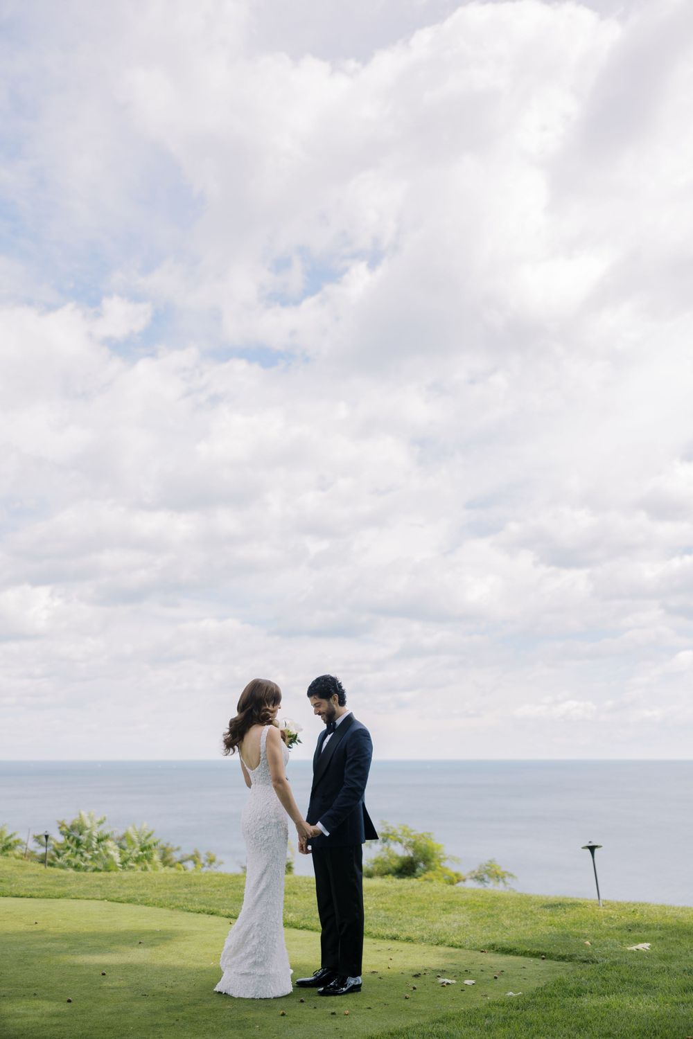 A couple in wedding attire stands on a hilltop overlooking the ocean under a cloudy sky.