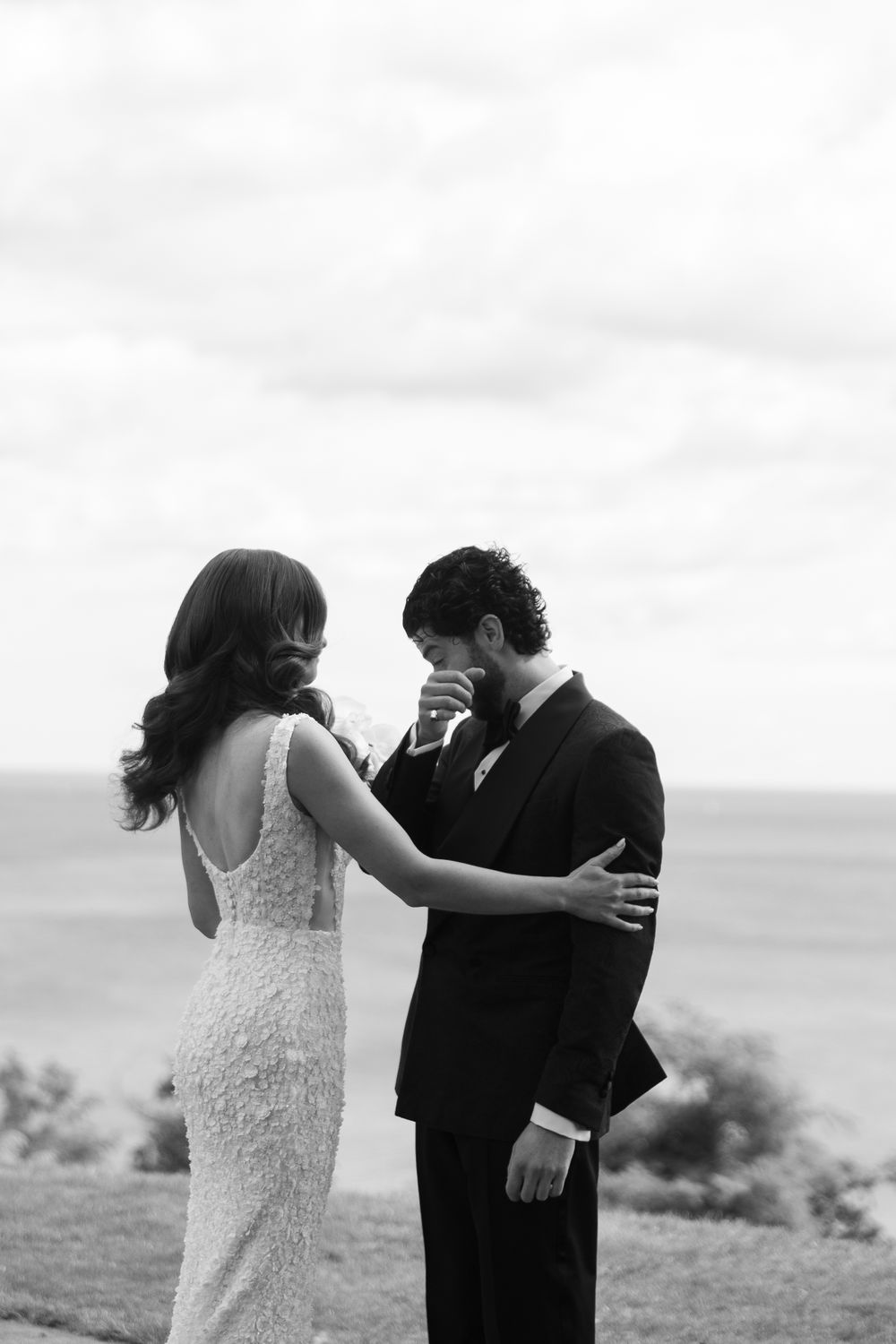 Black and white wedding photo of an emotional first look moment between a bride in lace dress and groom outdoors.
