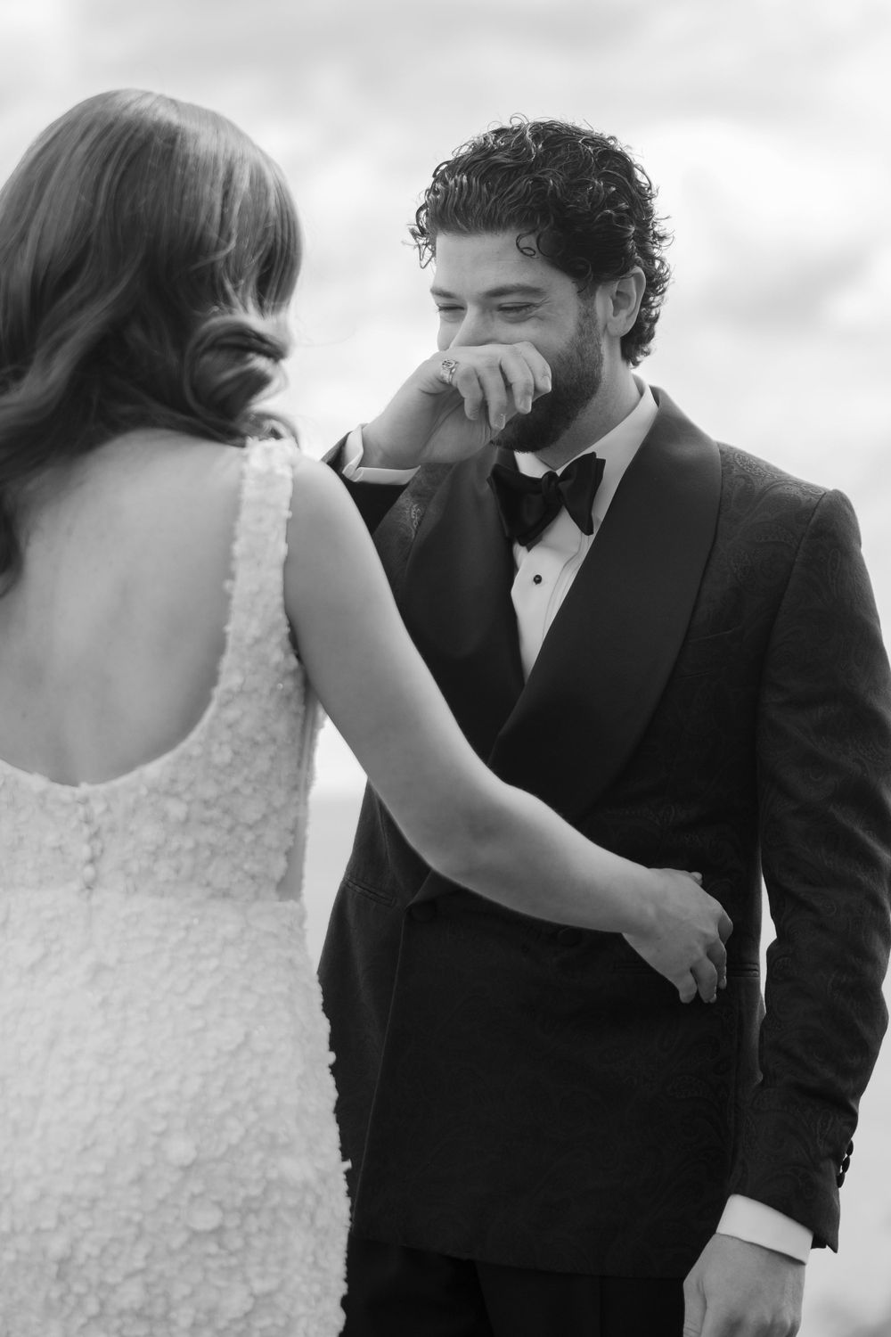 A black and white wedding photo shows an emotional moment between a bride in a lace dress and groom in a tuxedo.