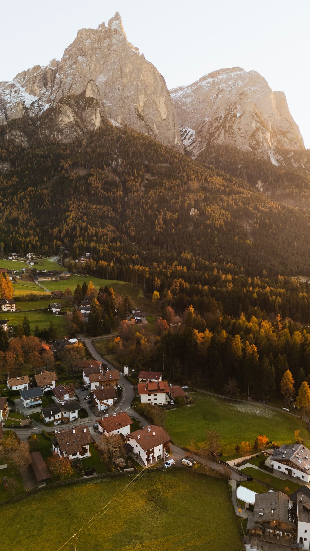 Aerial panorama of a charming alpine village nestled beneath towering mountain peaks with autumn foliage in the Dolomites.