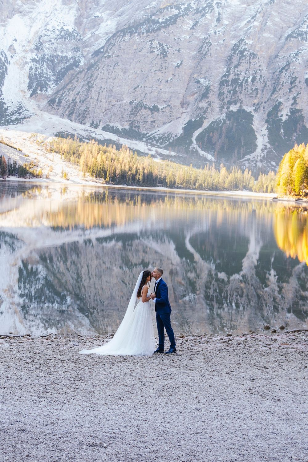 Couple embraces by tranquil mountain lake with autumn trees reflecting in calm waters at sunset.