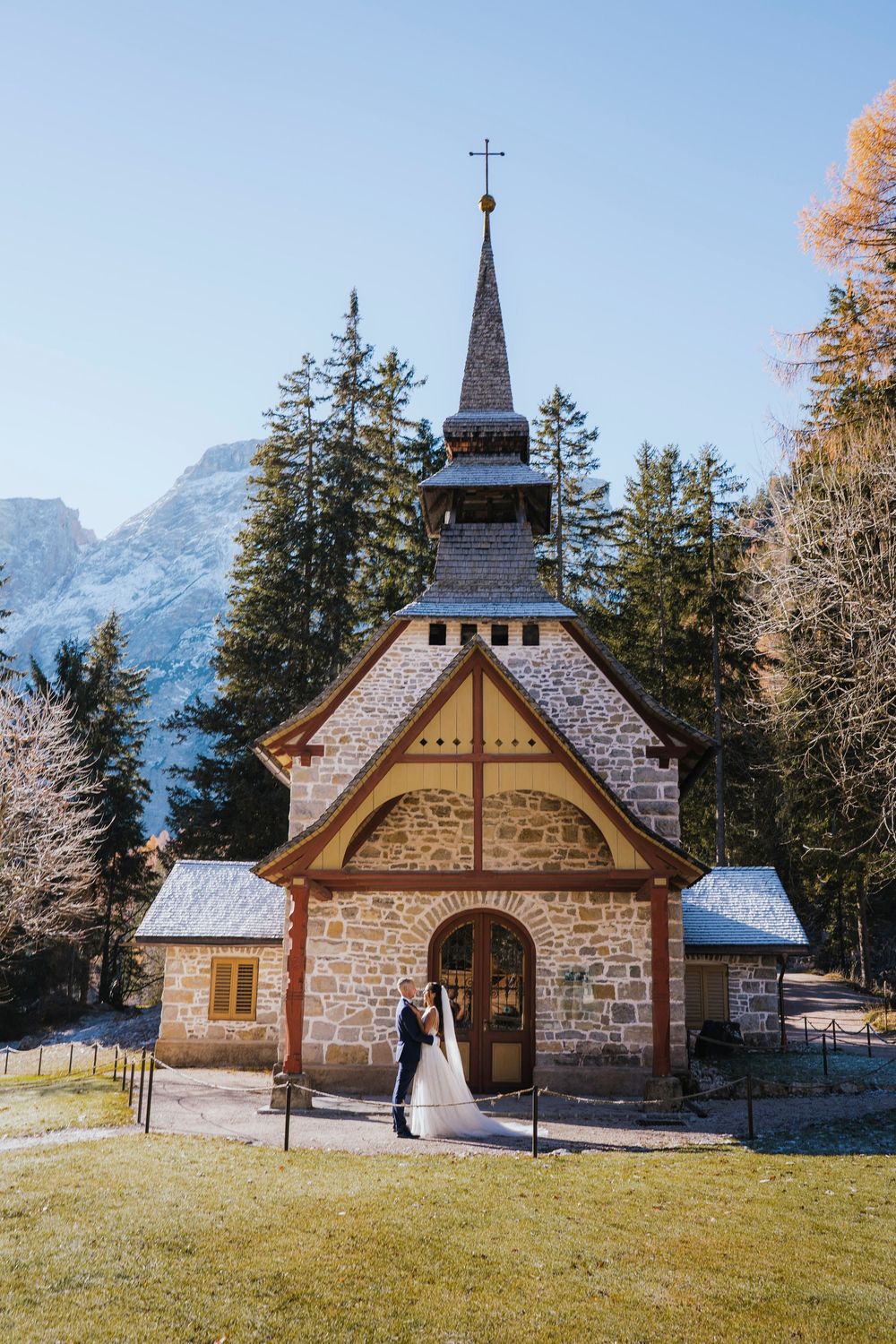 A charming stone chapel with a tall spire nestled in the Dolomite mountains surrounded by evergreen trees.