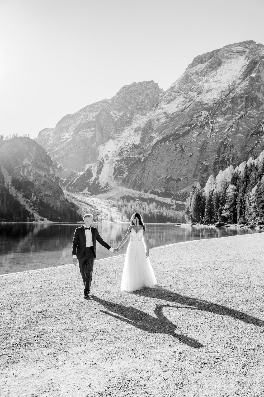 A couple walks along a scenic mountain lake shore with dramatic peaks rising in the background.