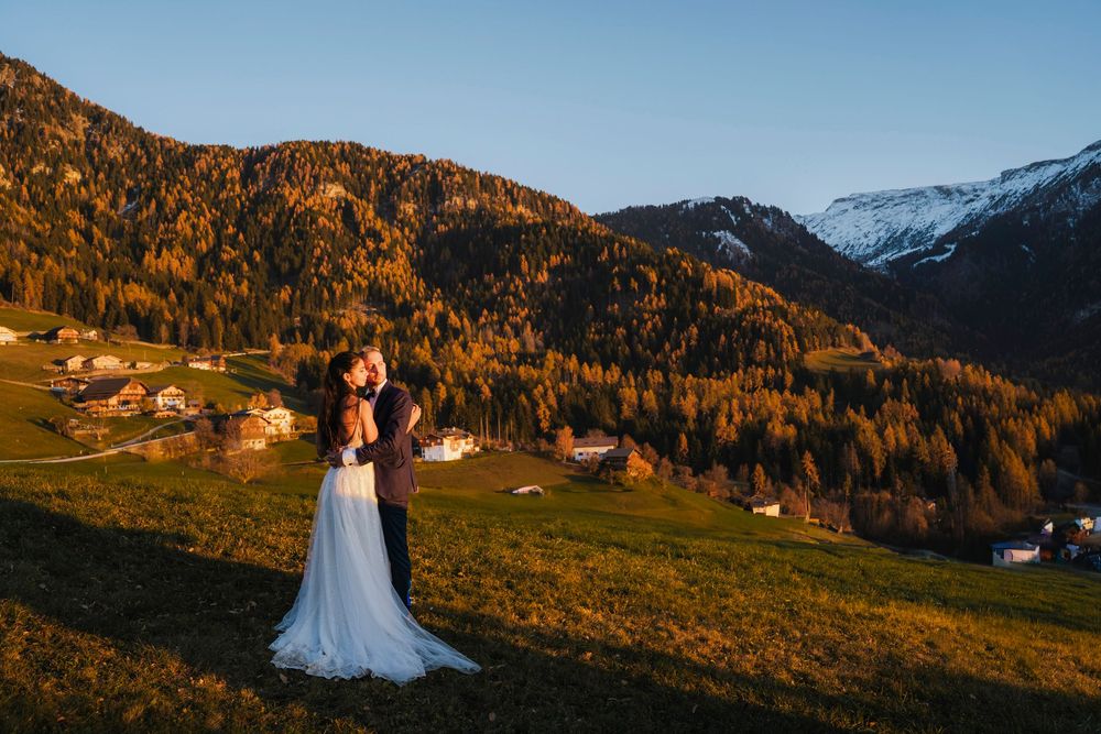 A couple in wedding attire poses in front of snow-capped mountains during golden hour in the Alps.