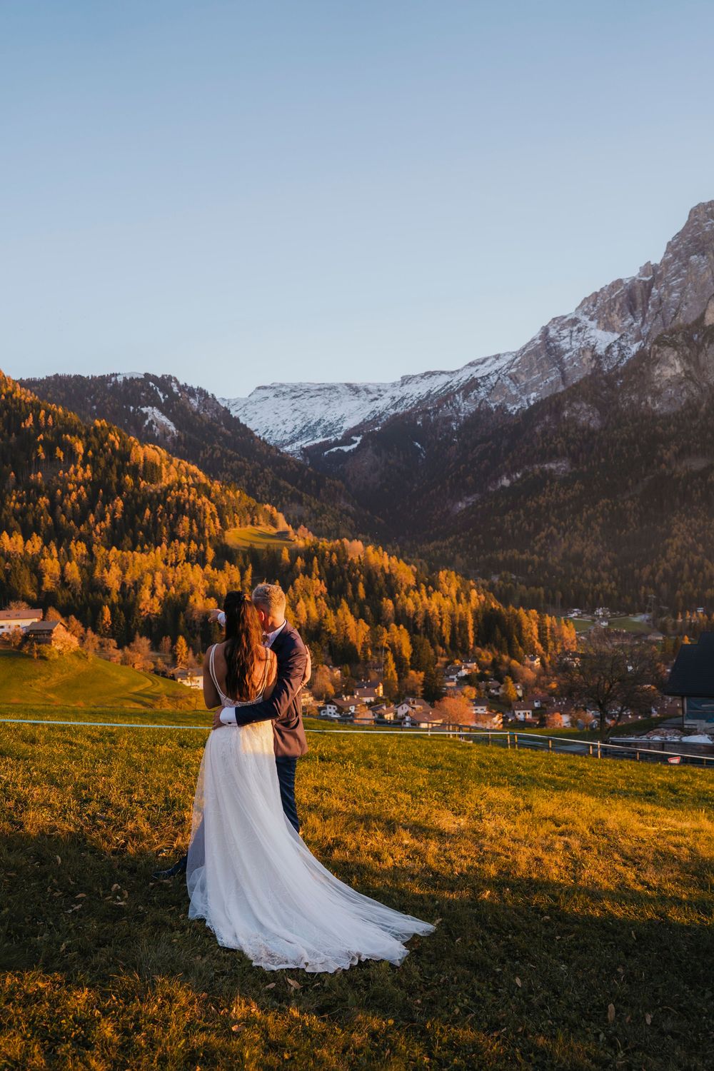 A couple embraces on a grassy hillside overlooking an alpine village with golden autumn trees.