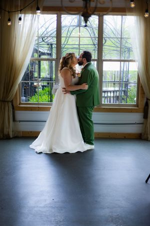 Wedding couple in green suit and white dress dancing in a sunlit room with floor-to-ceiling windows.