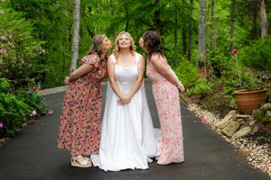 Three women in floral and white dresses share a candid moment on a garden path surrounded by lush greenery.