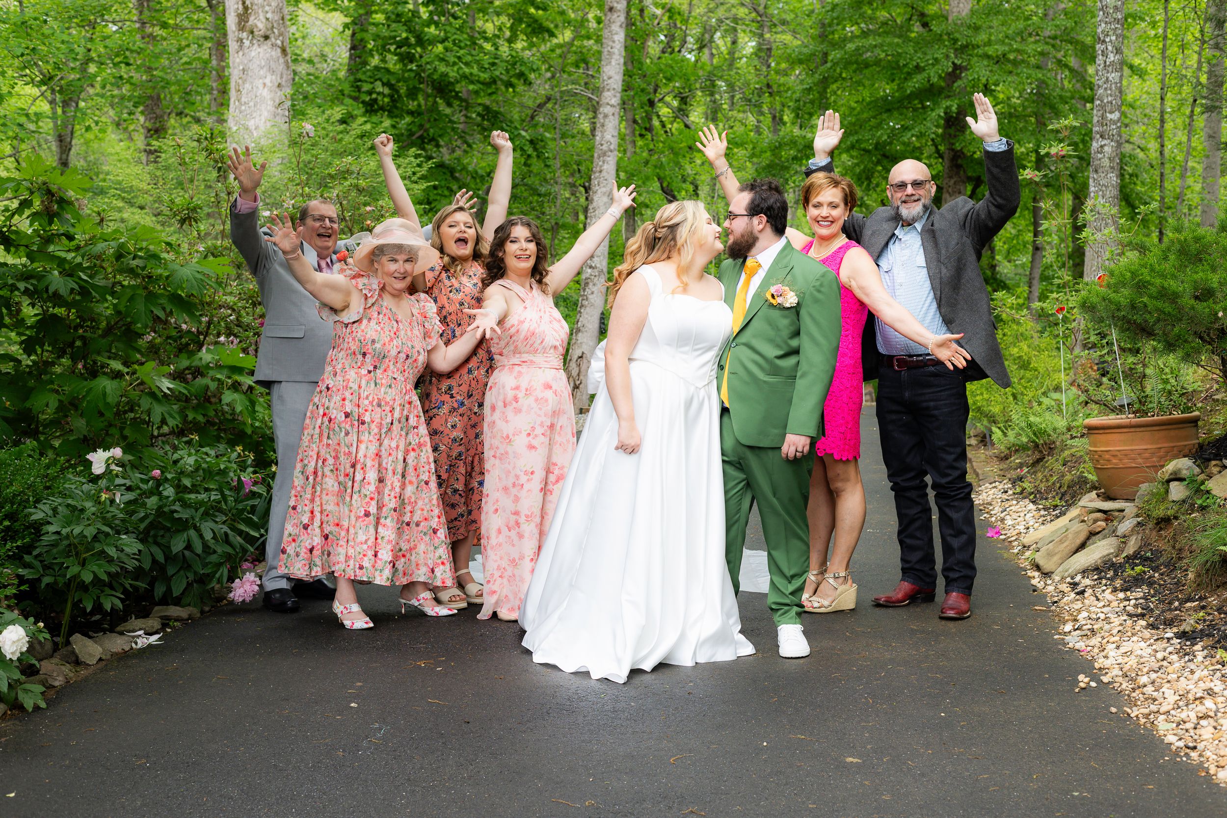 A joyful wedding party celebrates on a tree-lined pathway with bridesmaids in floral dresses raising their arms in excitement.