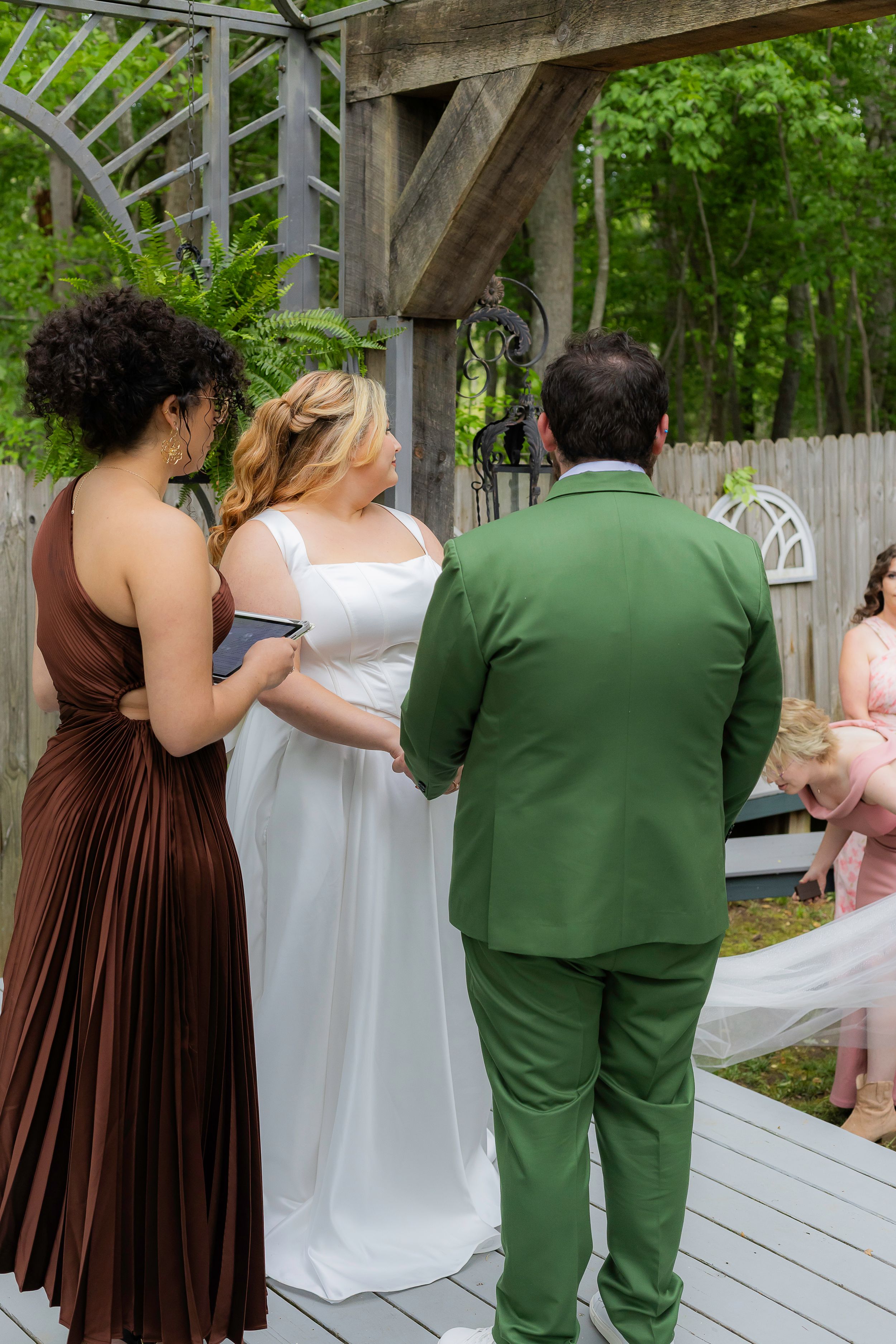 Wedding party on wooden deck with bridesmaids in brown dresses and couple in green suit and white gown.