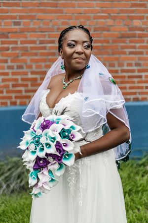 Bride in white dress holds stunning teal and purple calla lily bouquet against brick wall.