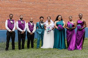 Wedding party poses outdoors against brick wall wearing teal and purple formal attire.