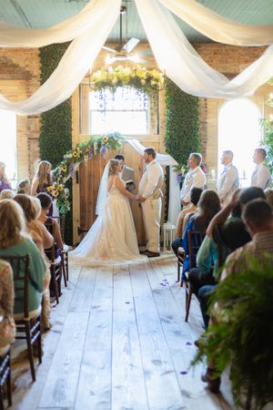 A wedding ceremony takes place in an elegant indoor venue with white draping and floral decorations.