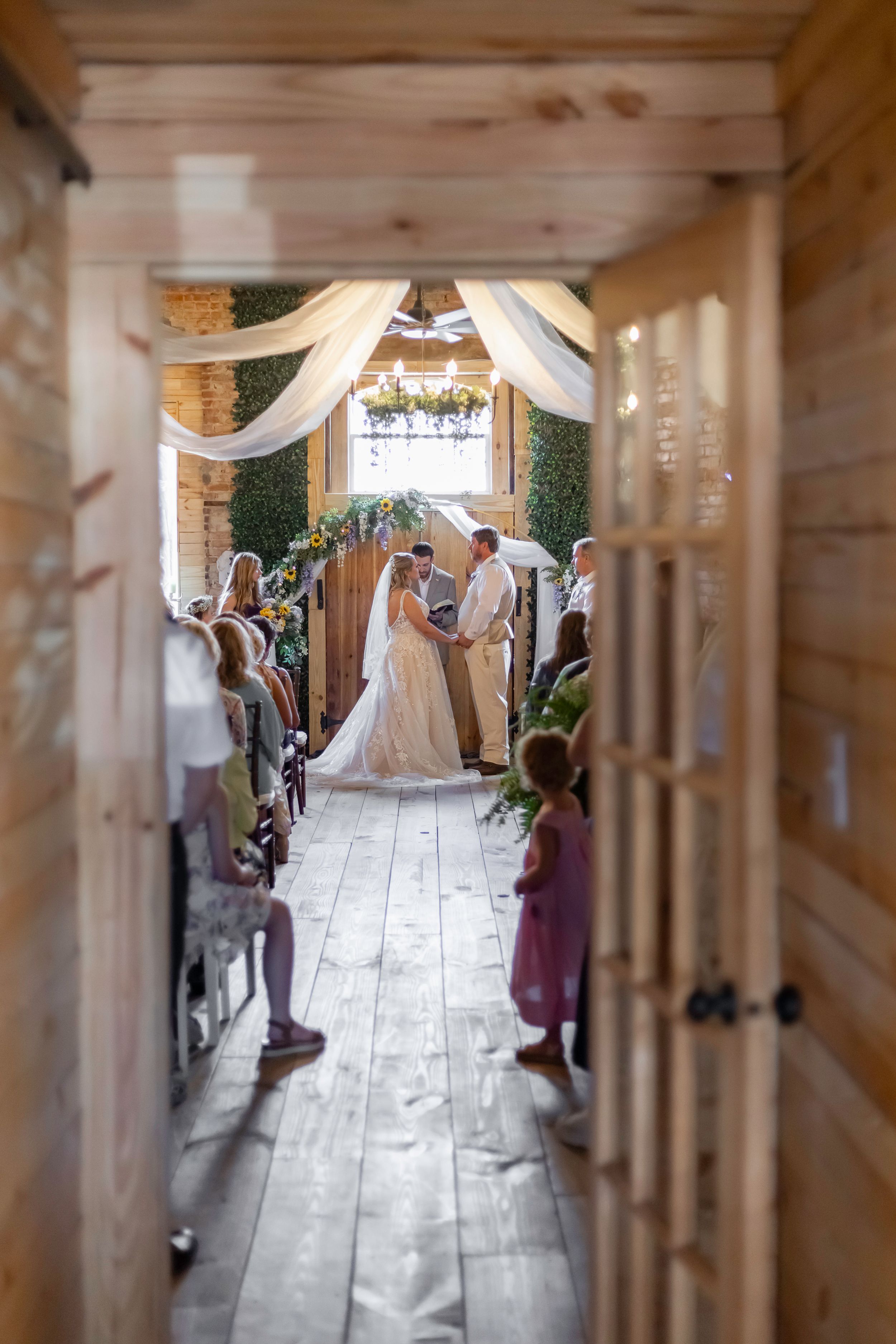 Wedding guests seated in rows watch a ceremony in a rustic wooden venue with white fabric decor.