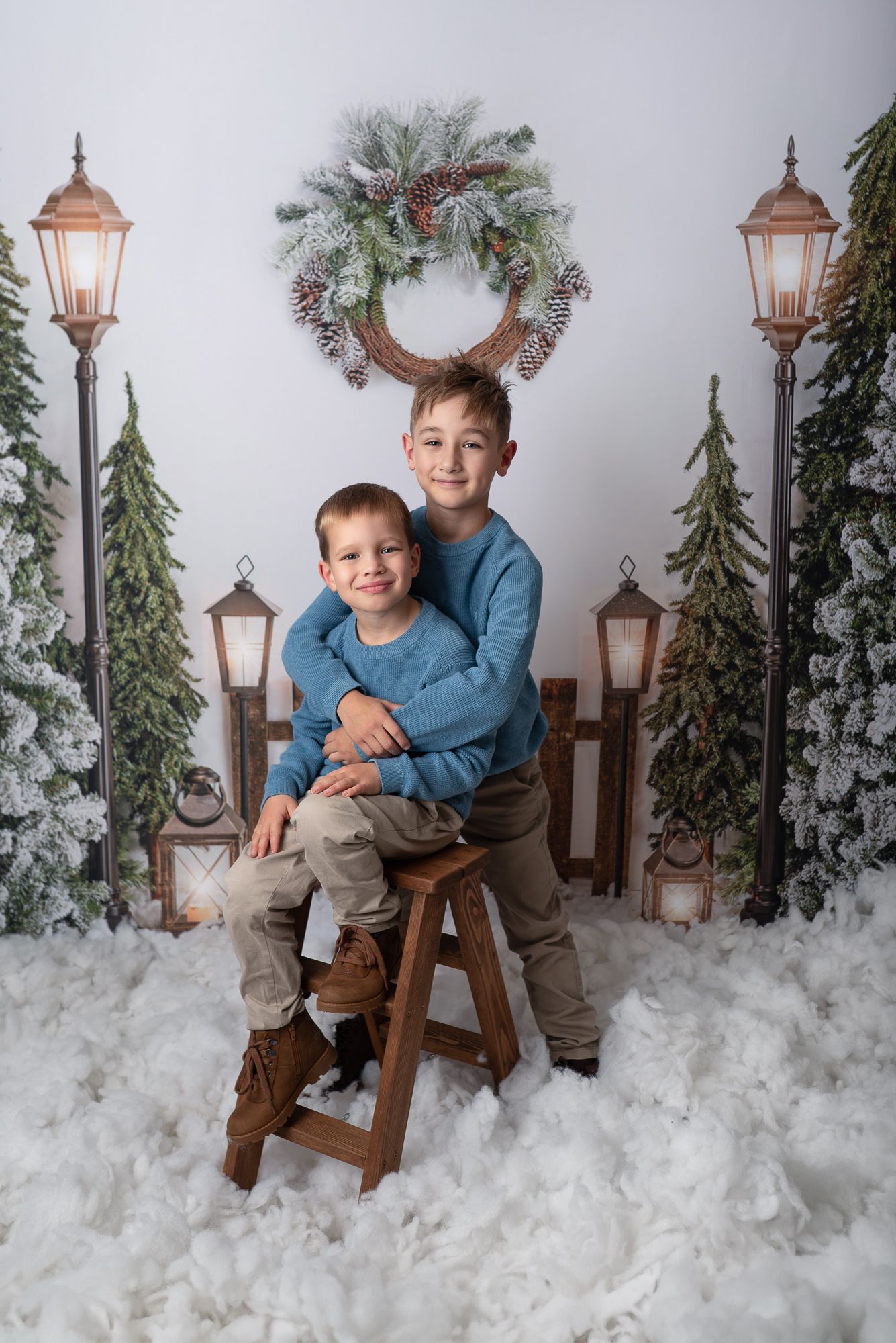 Festive winter portrait scene with decorative lamp posts, evergreen trees, snow, and holiday wreath against gray backdrop.
