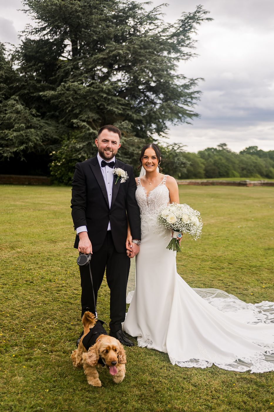 Newlyweds pose with their pet dog in countryside setting.