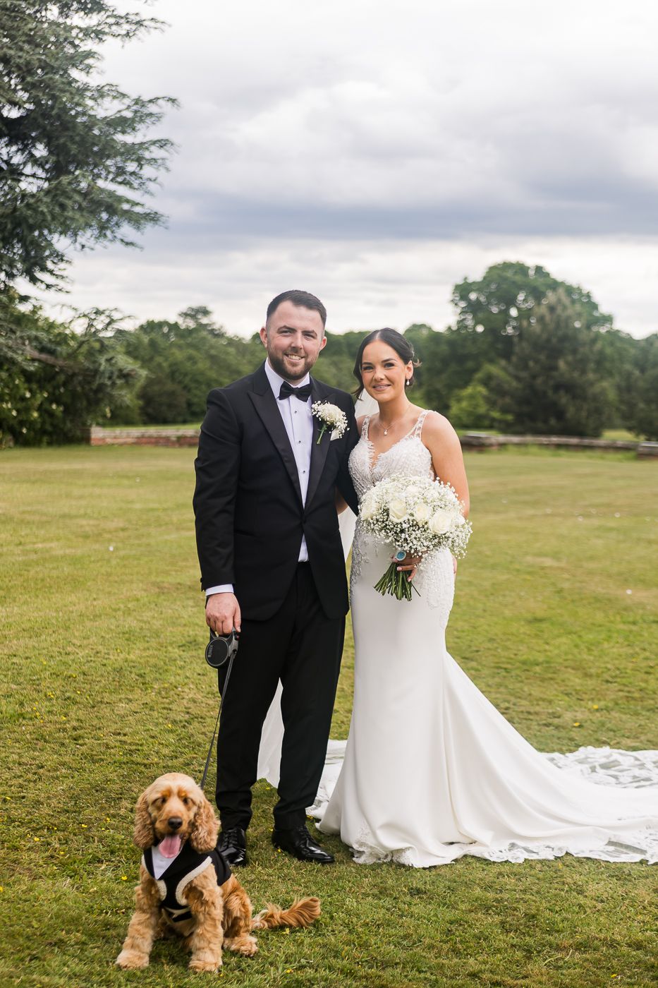 Wedding couple poses with their brown dog on a grassy field with cloudy sky backdrop.