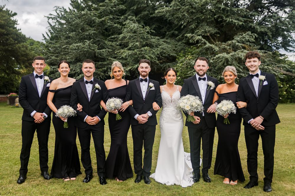 Wedding party in black attire standing in line on green lawn with evergreen trees.