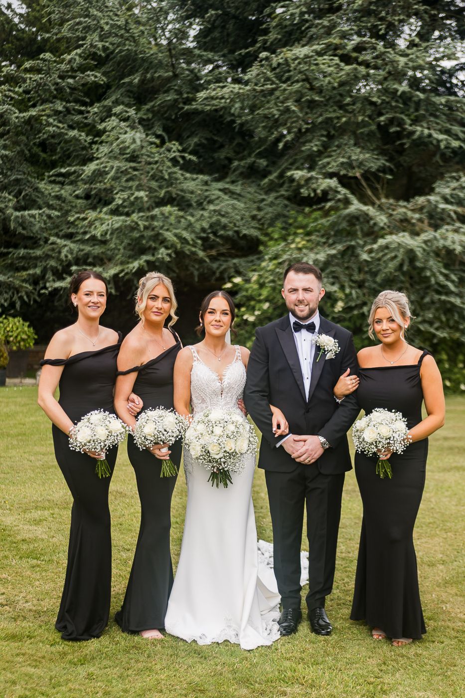 Wedding party in formal attire posing playfully on grass with evergreen backdrop.