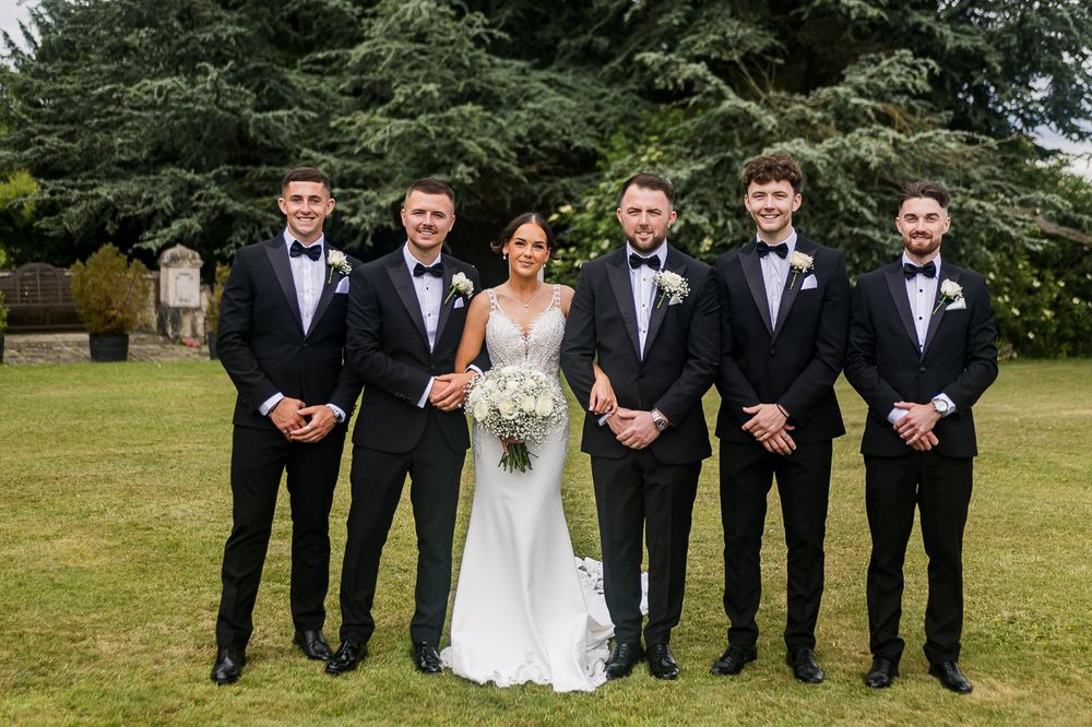 Formal wedding party lined up outdoors with evergreen trees in background.