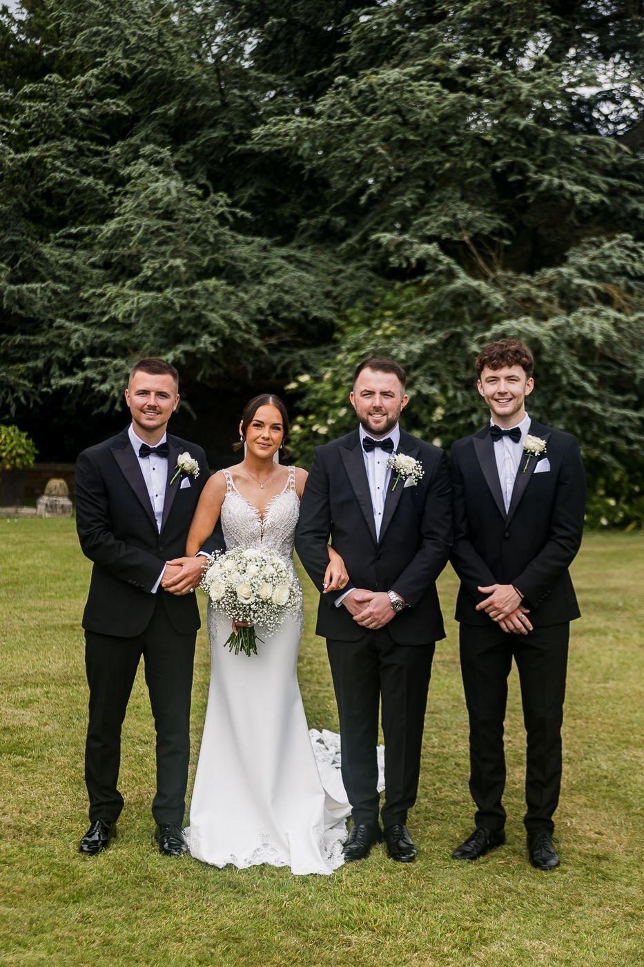 Wedding party in formal attire posing casually on grass with trees behind.