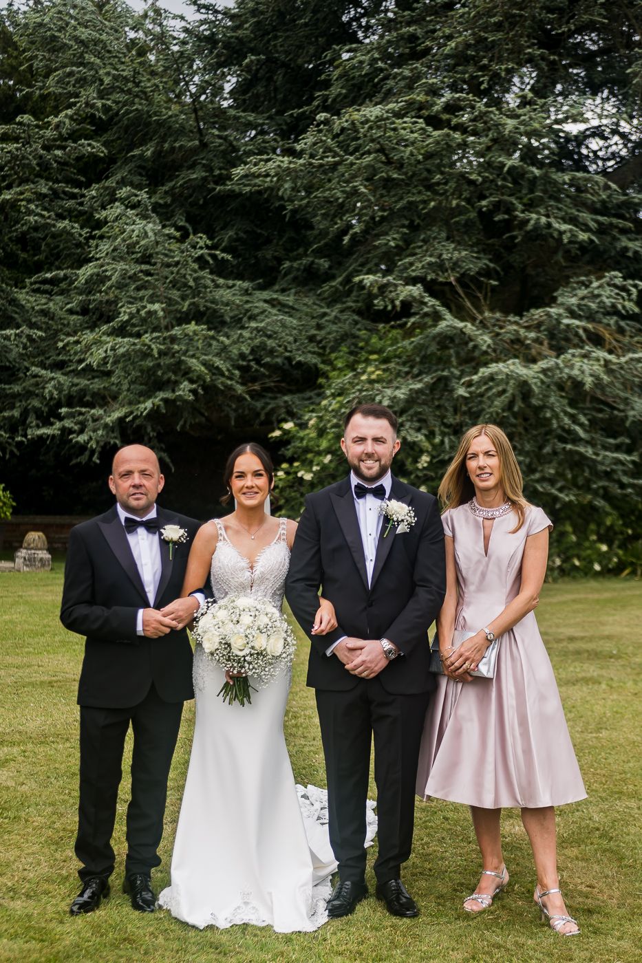 Wedding party in mixed formal attire posing together on grass with evergreen backdrop.