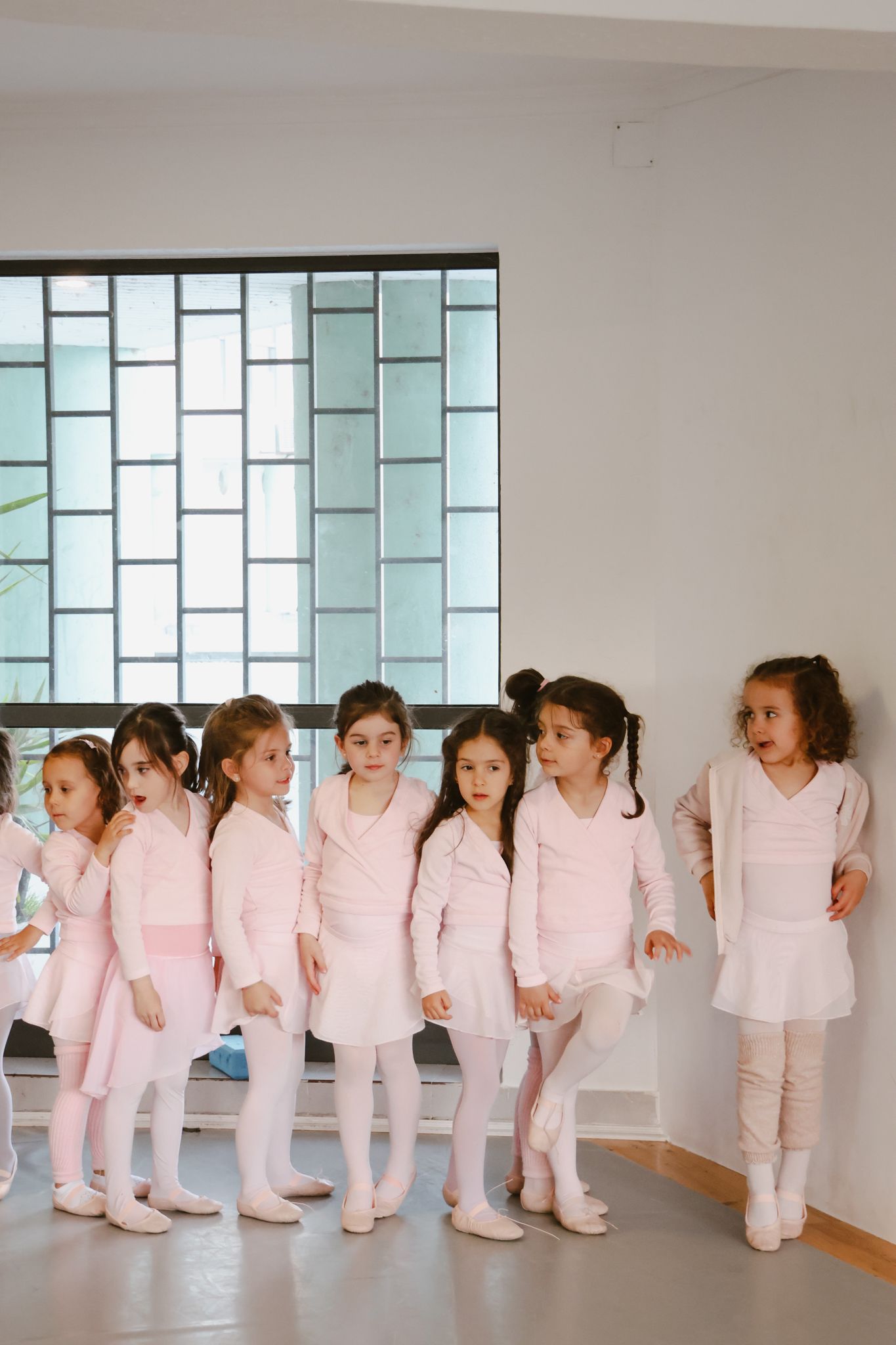Young ballet students in pink outfits lined up against a wall with geometric windows.
