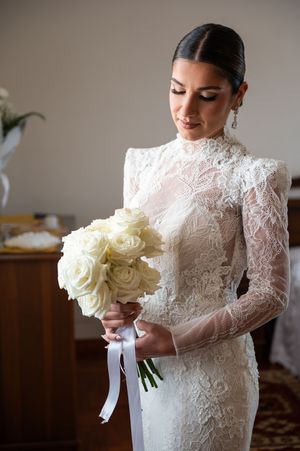 Bride in long-sleeved lace gown holding a classic white rose bouquet.