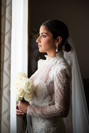 Bride in vintage-inspired lace gown poses with white roses near window light.