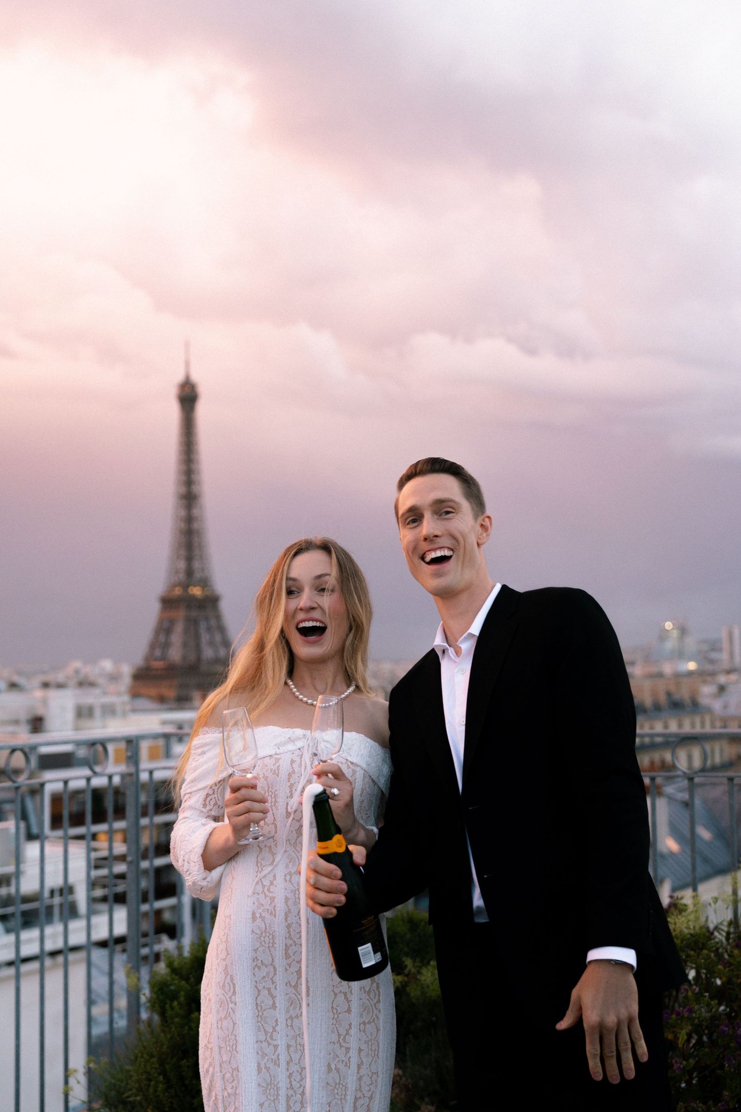 A couple shares laughter and drinks on a rooftop terrace with the iconic Eiffel Tower visible in the Paris skyline.