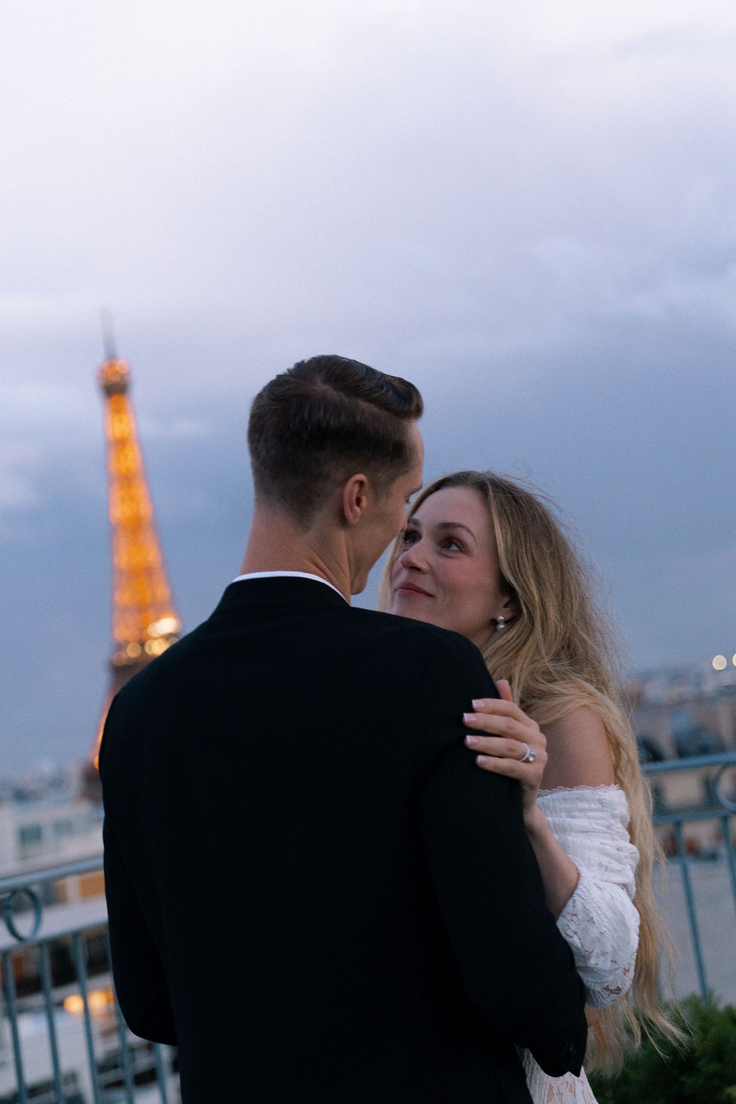 A couple embraces on a Parisian rooftop with the illuminated Eiffel Tower glowing in the evening background.