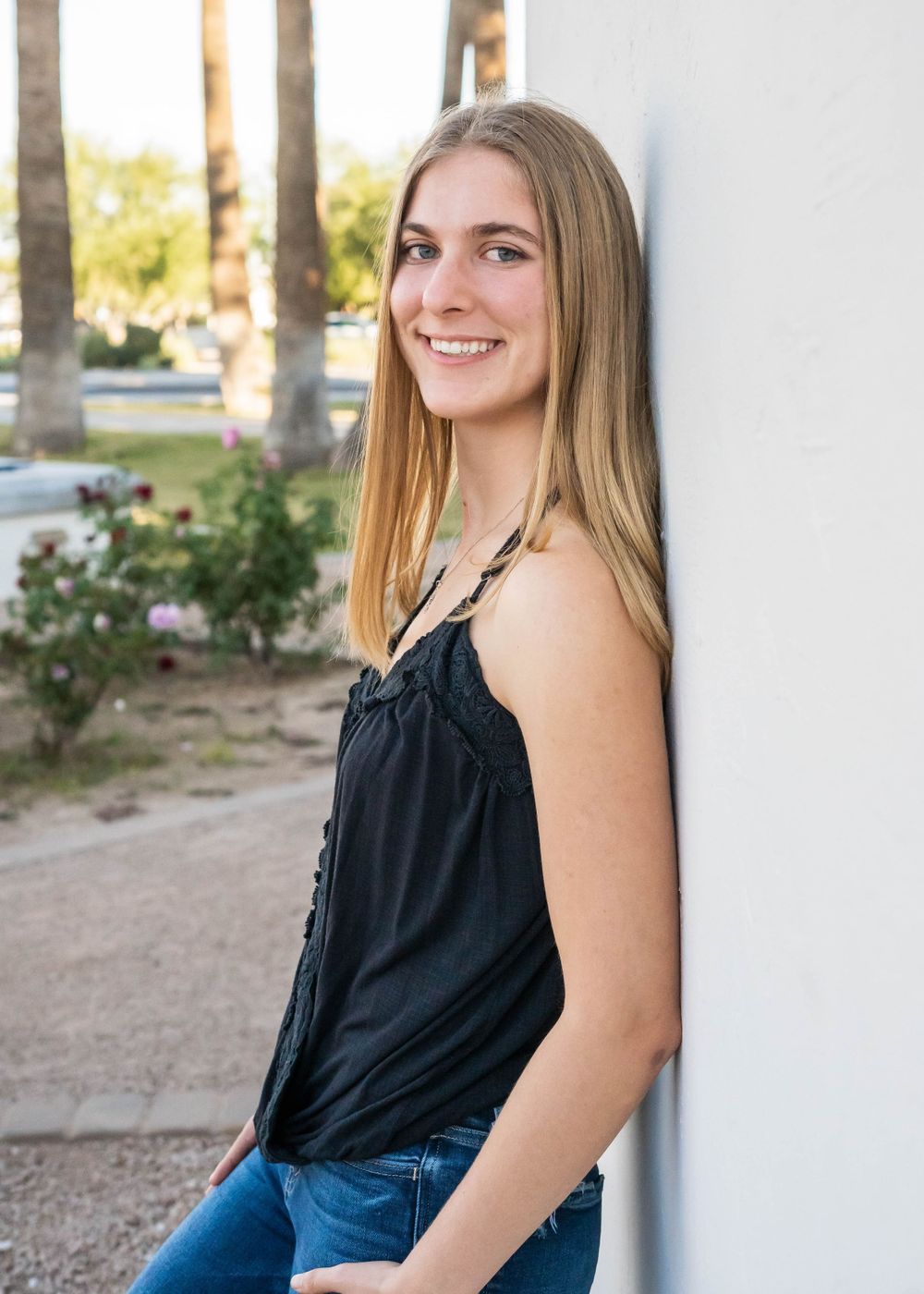 Woman in black tank top and jeans leaning against white wall with palm trees in background.