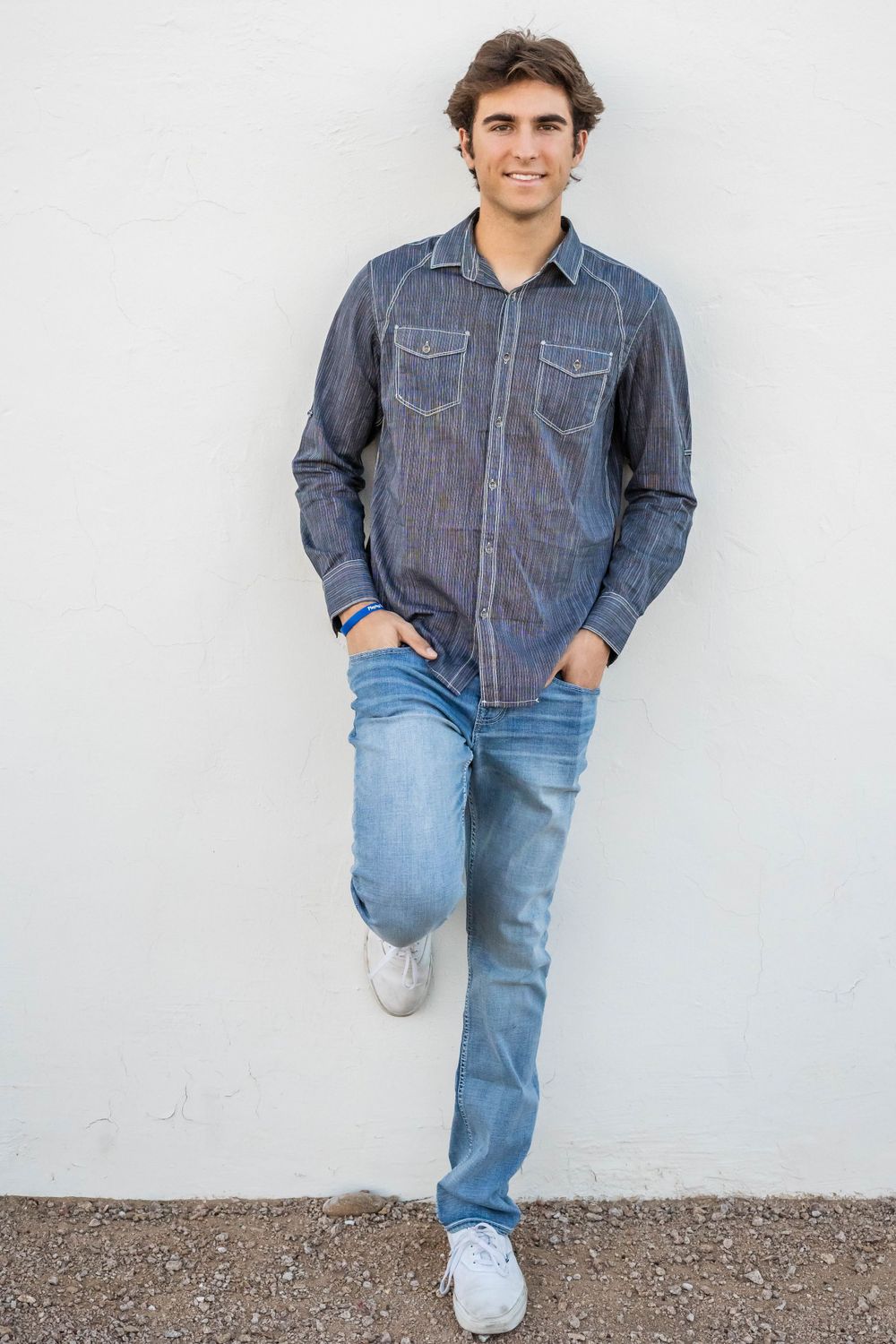 A casual portrait of a person in denim shirt and jeans leaning against a white wall.