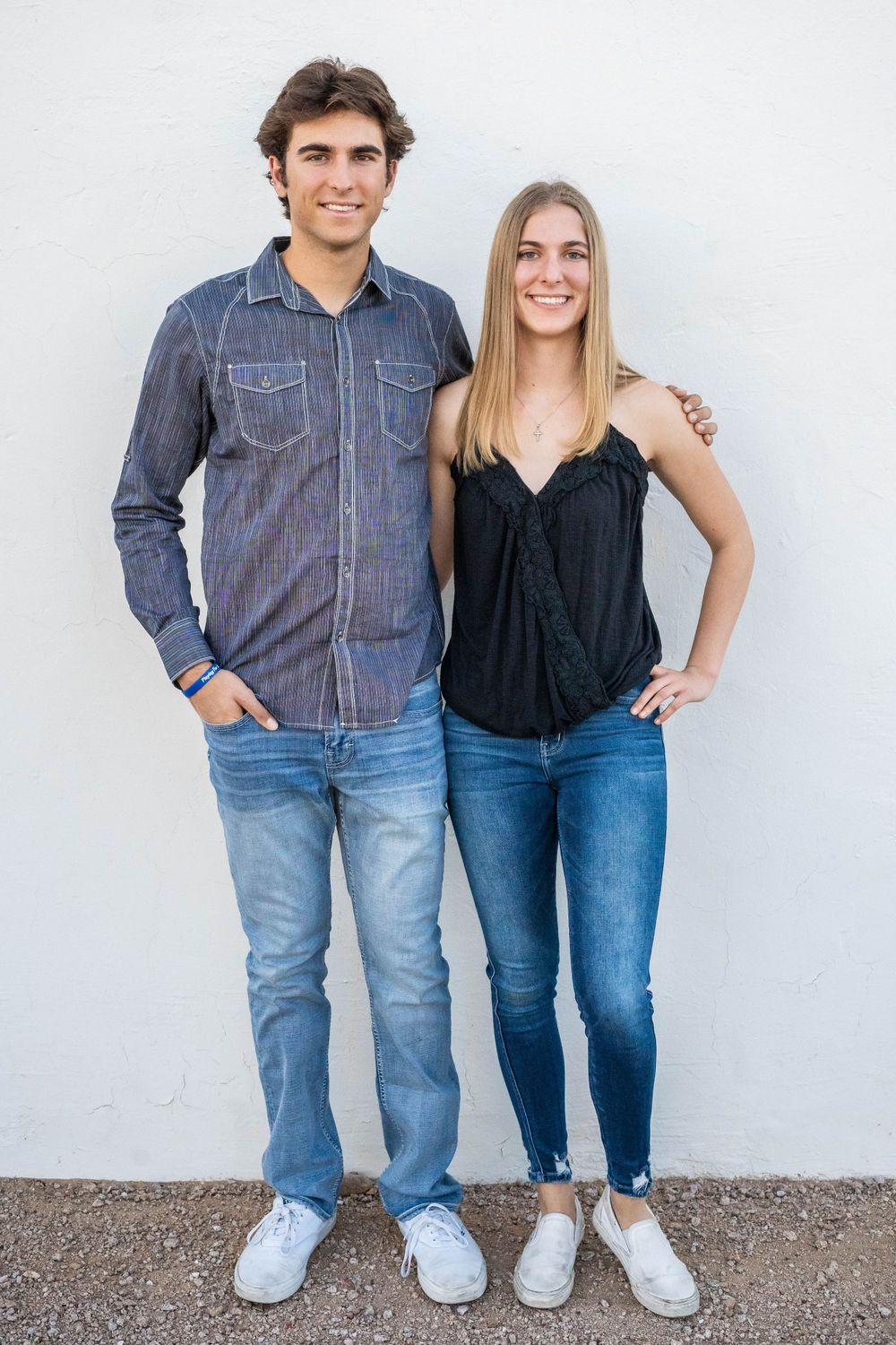 Casual couple in denim outfits standing against white wall wearing matching blue jeans and sneakers.