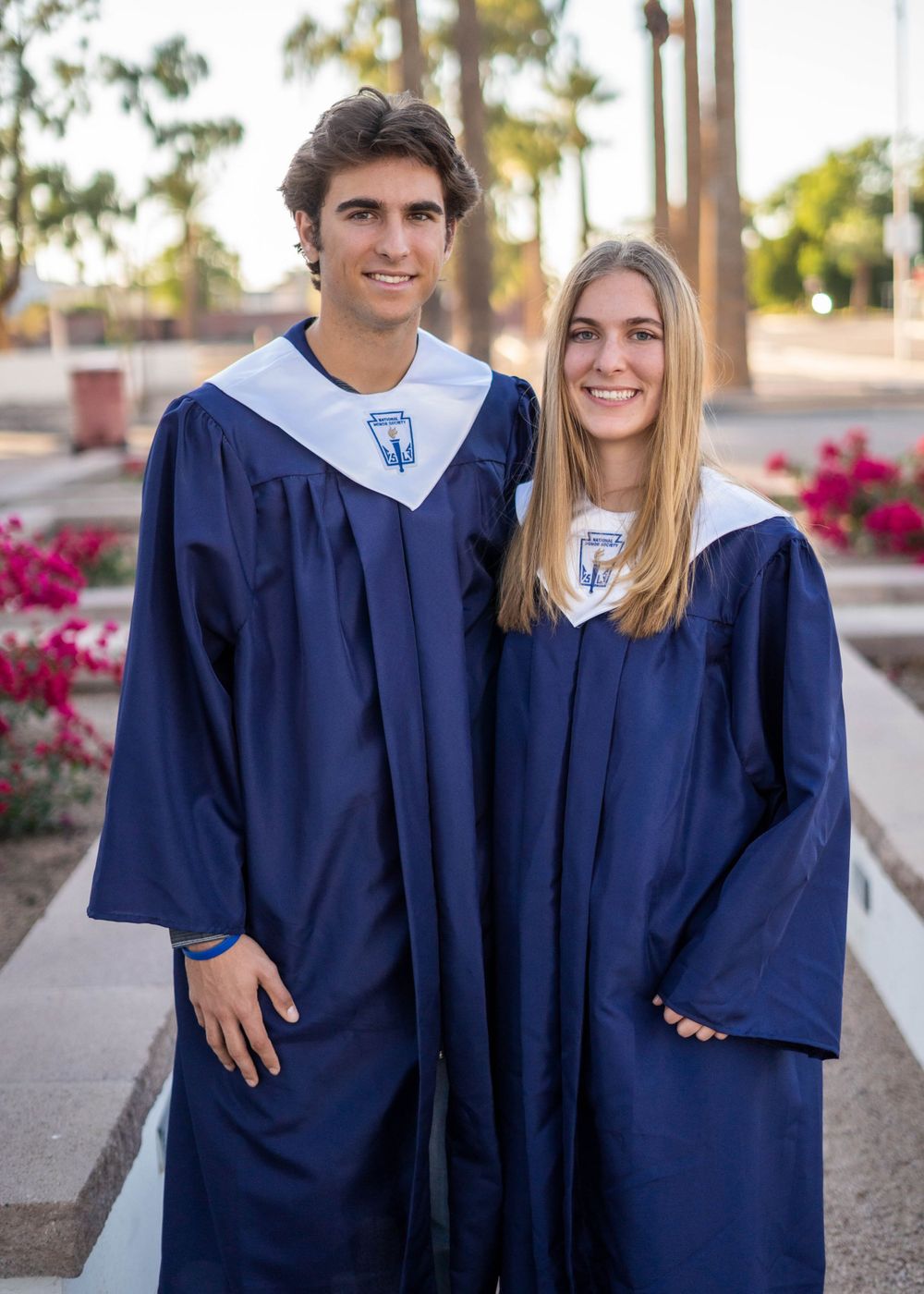 Two graduates pose in blue academic gowns with matching stoles on a campus pathway.