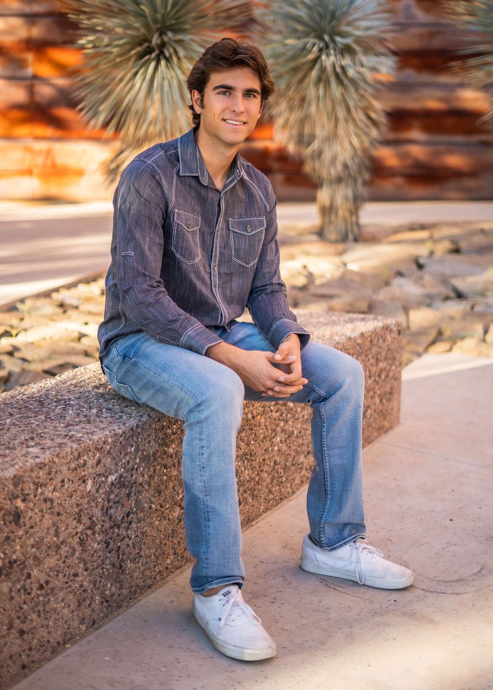 Man in denim shirt sitting on stone ledge with desert plants and warm sunset lighting.