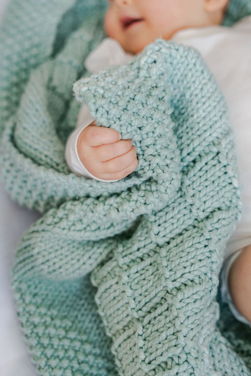 Newborn baby wrapped in a soft mint green knitted blanket, tiny hand visible against the cozy texture.