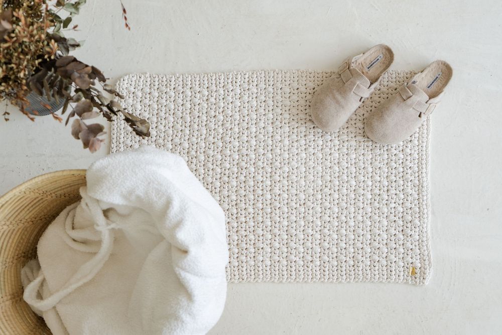 Neutral toned crochet bathmat styled with beige shoes, a straw hat, and white linen on a light surface.
